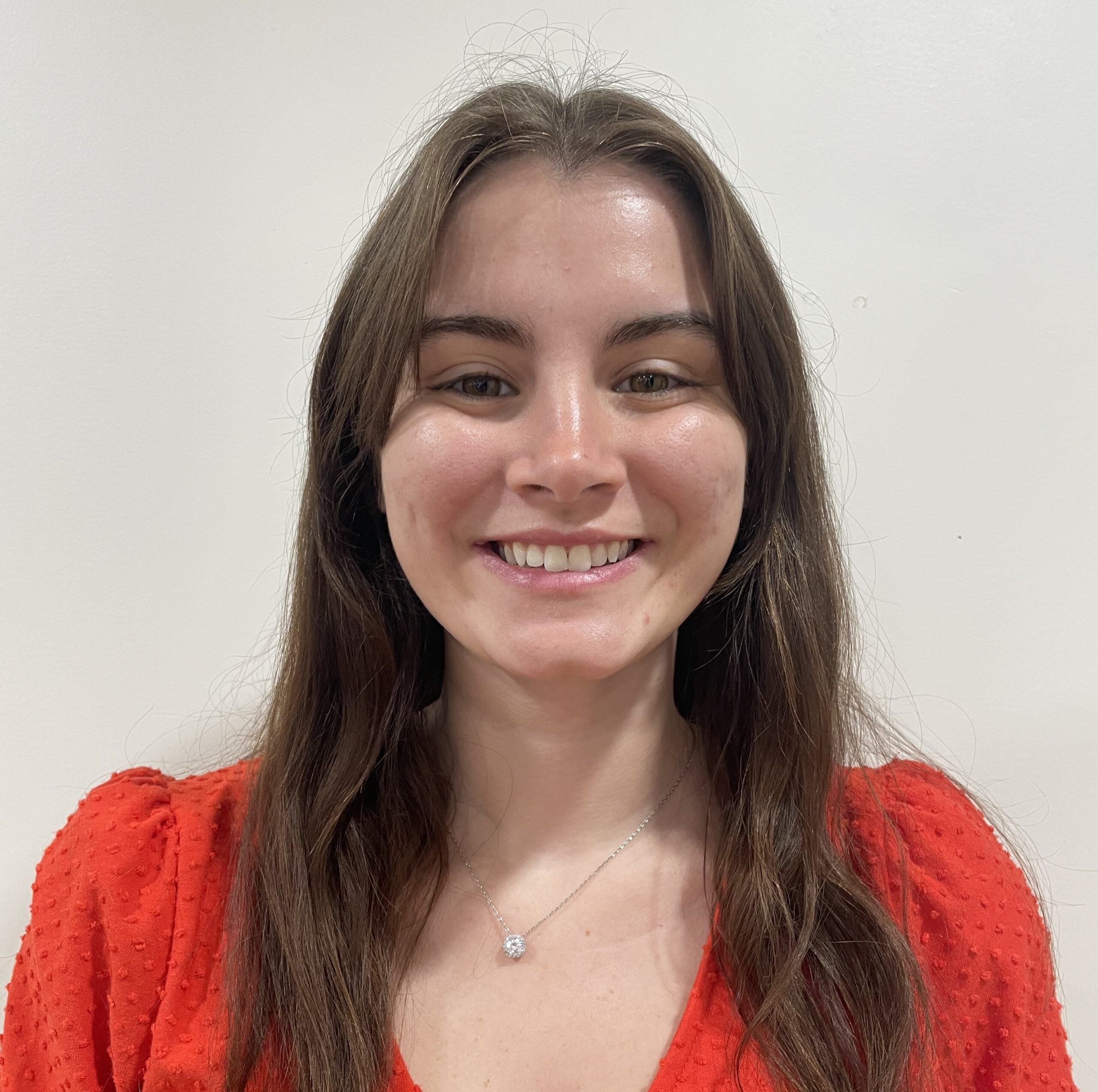 A smiling young woman with long brown hair stands against a plain light background, wearing a bright red short-sleeved V-neck textured top and a small round pendant necklace.