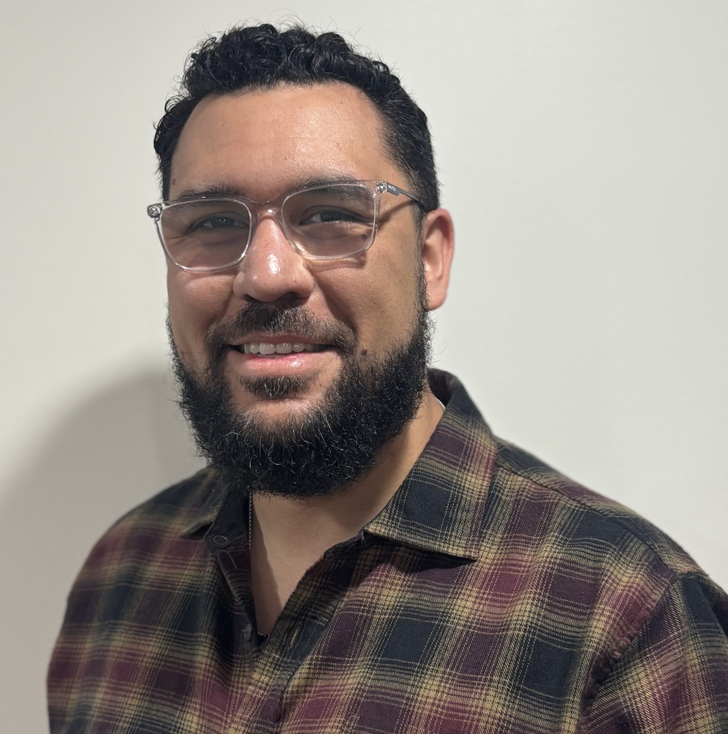 A smiling man with short curly dark hair and a full beard stands against a plain light background, wearing clear-framed glasses and a dark plaid button-up shirt.