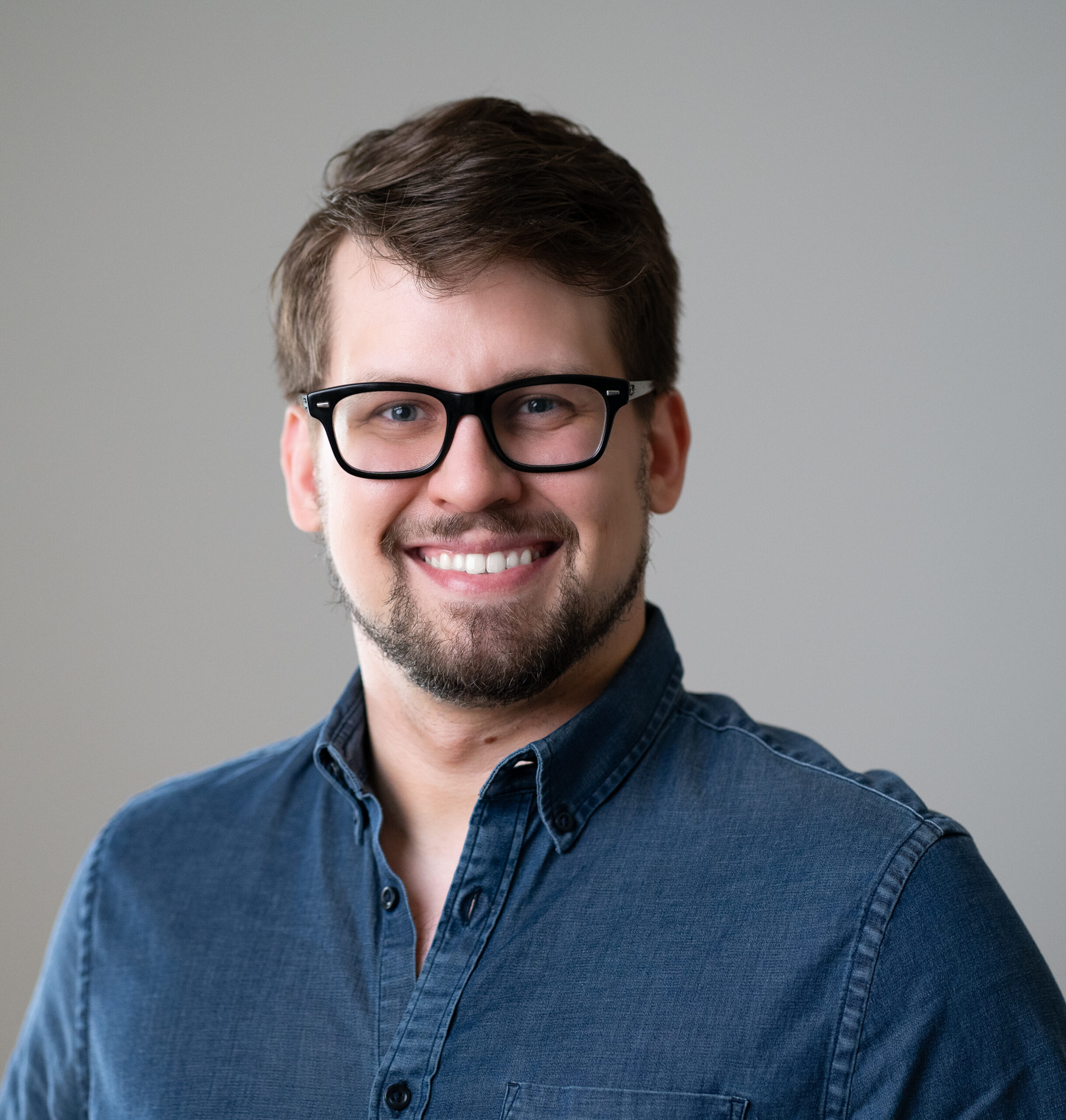 Portrait of a smiling person with short brown hair, black rectangular glasses, and a trimmed beard, wearing a blue button-up shirt against a light gray background.