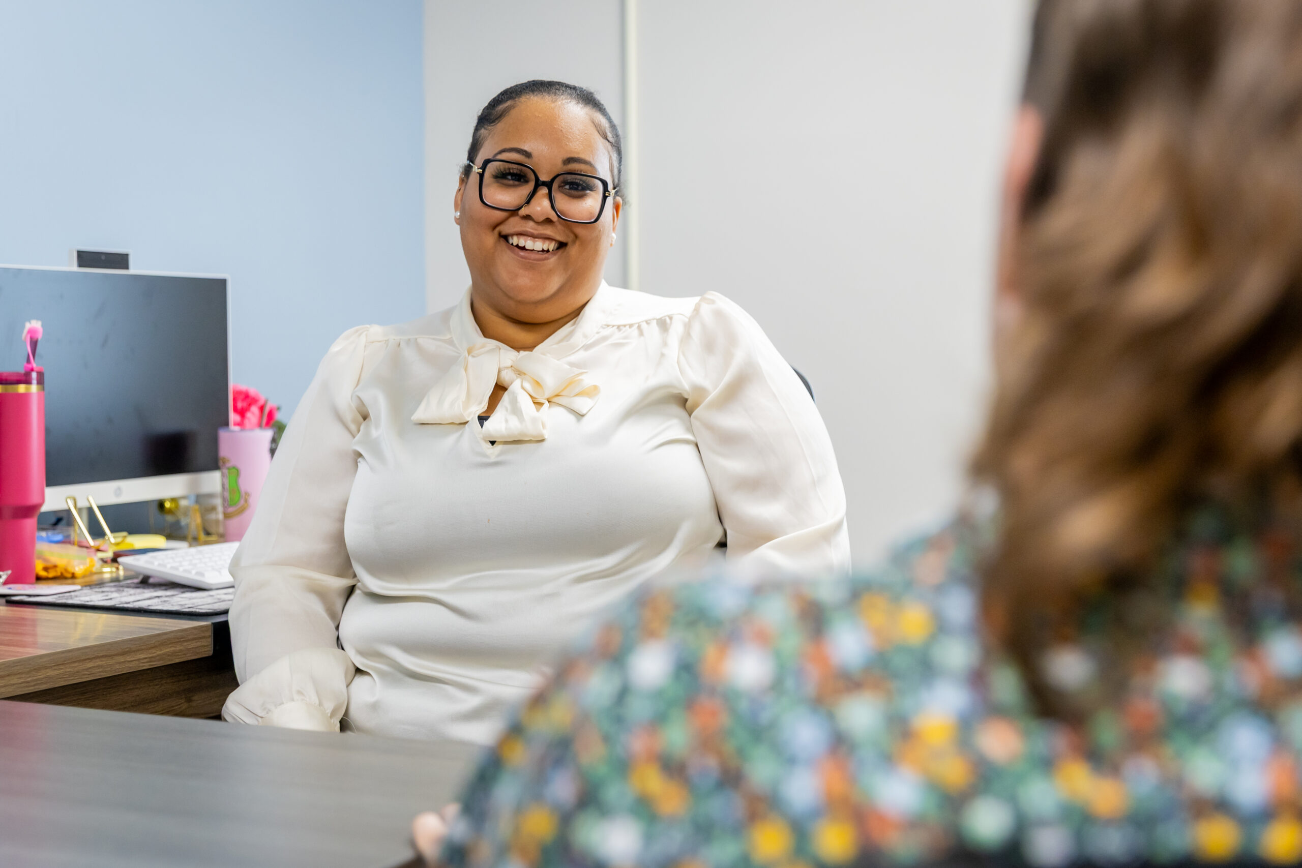 Therapist seated at a desk smiling during a counseling session, with a client blurred in the foreground in a bright office.
