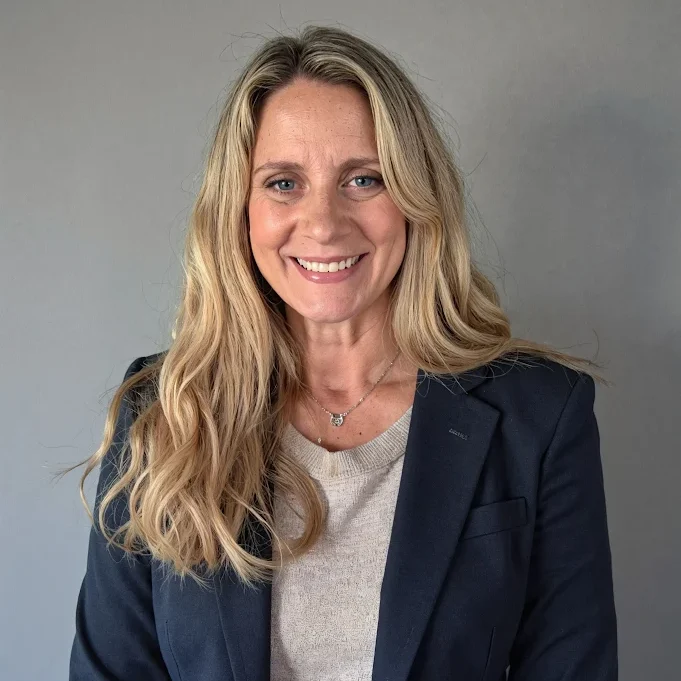 Professional headshot of a smiling woman with long blonde hair wearing a navy blazer and light sweater against a neutral gray background.